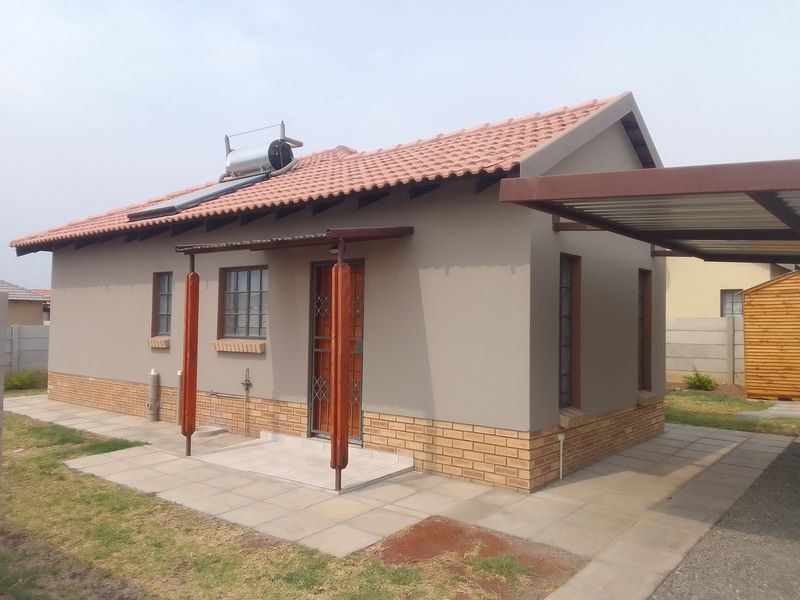 A well-maintained townhouse exterior featuring a tiled roof, solar water heater, brick base detailing, and an attached carport structure.