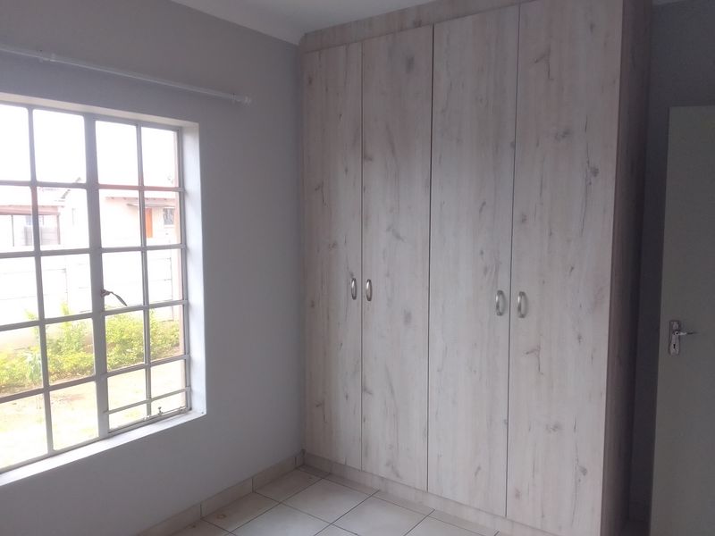A bright bedroom featuring light wood-grain built-in cupboards, tiled flooring, and a window with security bars overlooking the exterior.