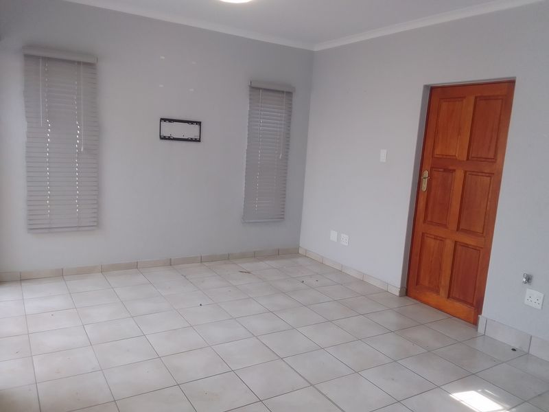 An empty living room featuring neutral tiled flooring, light grey walls, wooden door, and fitted venetian blinds.
