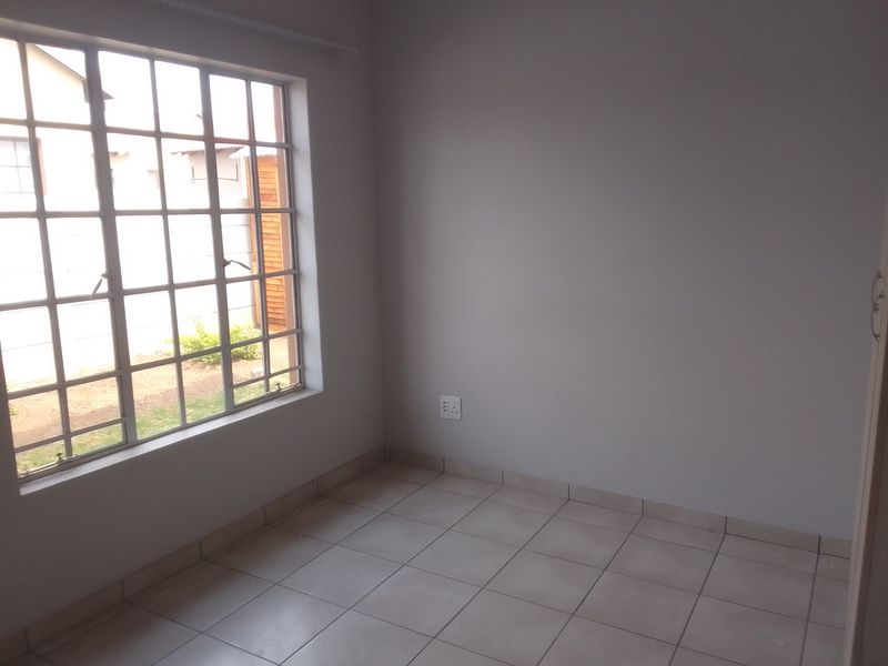 A bright, empty bedroom featuring neutral tiled flooring, a large window with security bars, and a clean, minimalist aesthetic.