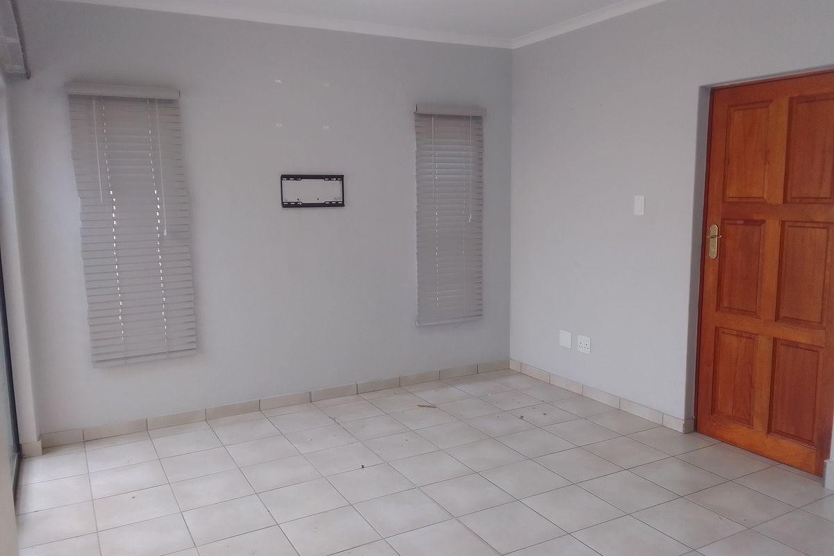 An empty living room featuring neutral tiled flooring, grey walls, venetian blinds, and a solid timber door.