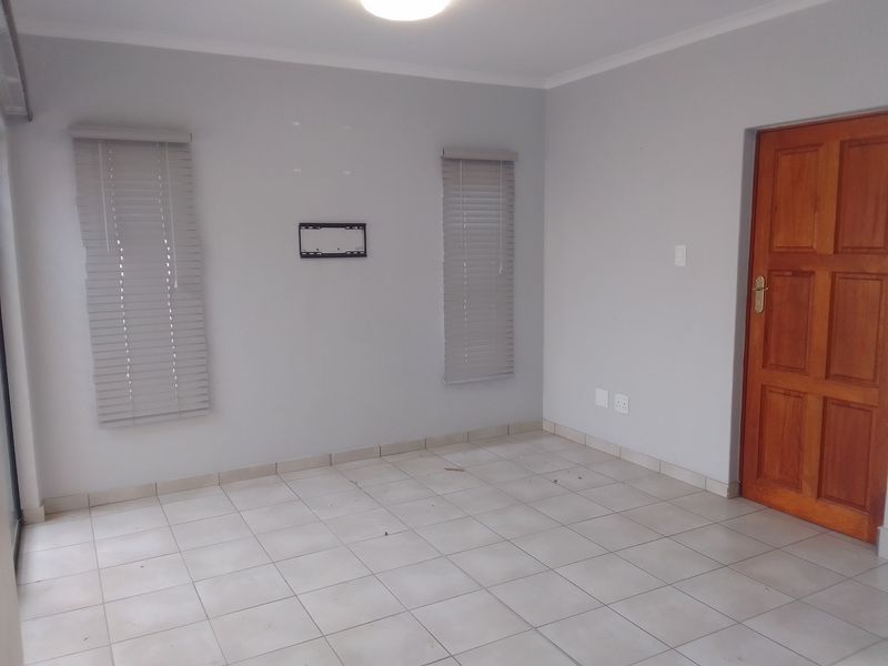 An empty living room featuring neutral tiled flooring, grey walls, venetian blinds, and a solid timber door.