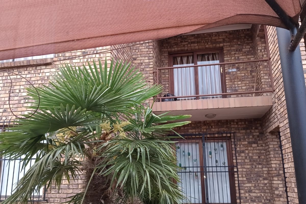 Rear view of a face brick townhouse featuring a private balcony with metal balustrades, security bars on windows, and a shade cloth covered patio area.