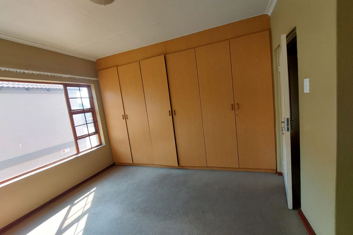 A spacious, empty bedroom featuring extensive built-in wooden cupboards, carpeted flooring, and a window with security bars.