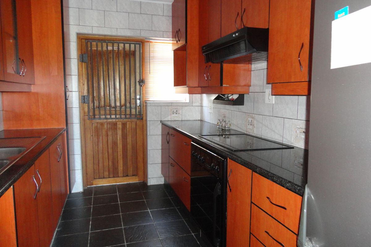 A functional galley-style kitchen featuring wood-finish cabinetry, dark granite countertops, a built-in hob and extractor fan, and tiled flooring with a security door.