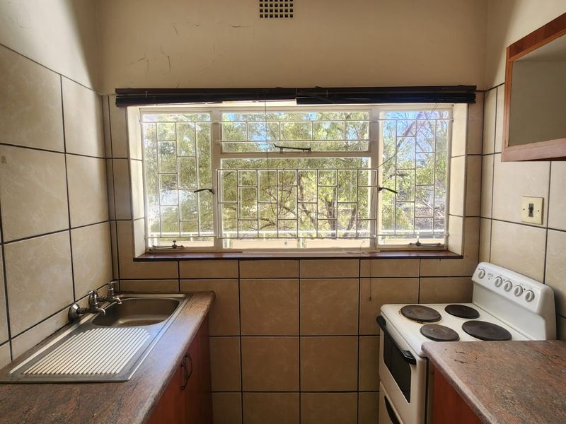 A compact kitchen featuring stone countertops, a stainless steel sink, and a freestanding stove, with tiled finishes and security bars on the window.