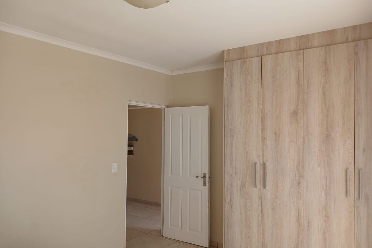 A bedroom featuring light-toned wood-grain built-in cupboards, neutral wall finishes, and tiled flooring, leading into an adjacent hallway.