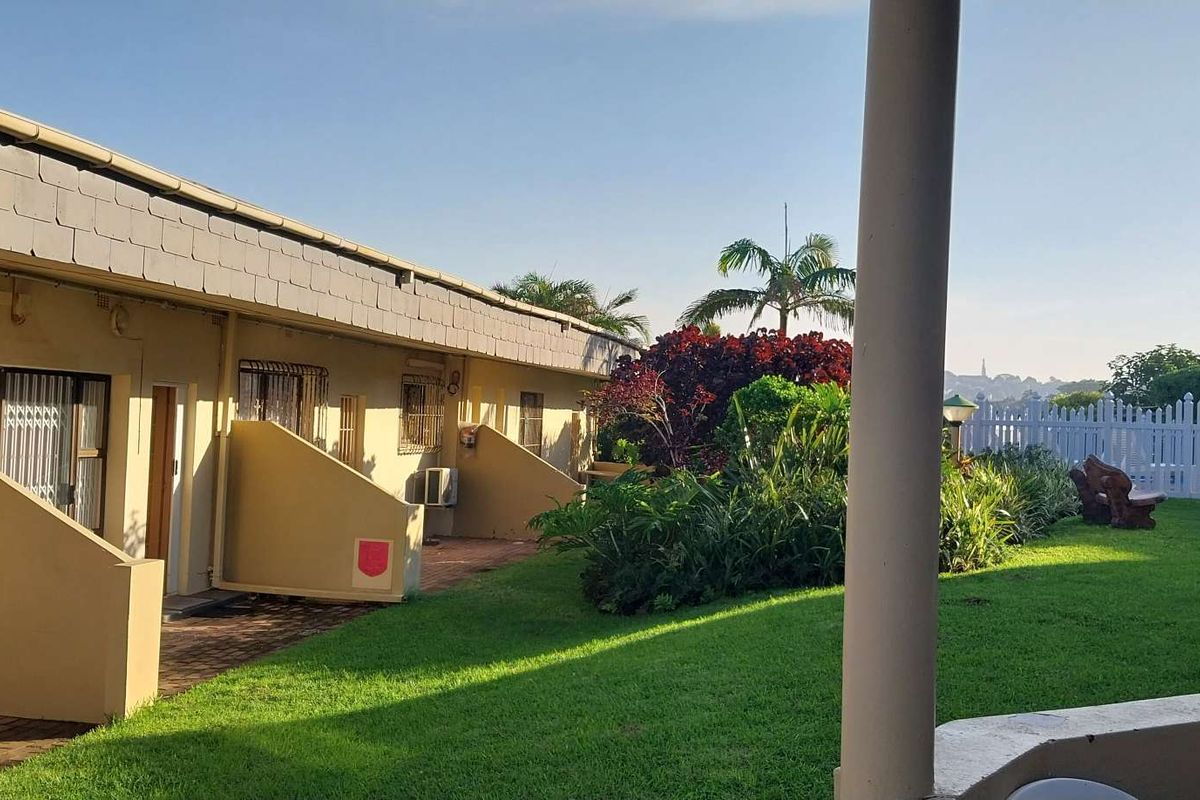 A view of the apartment exterior featuring a paved walkway, manicured lawn, and established garden beds with a white picket fence boundary.