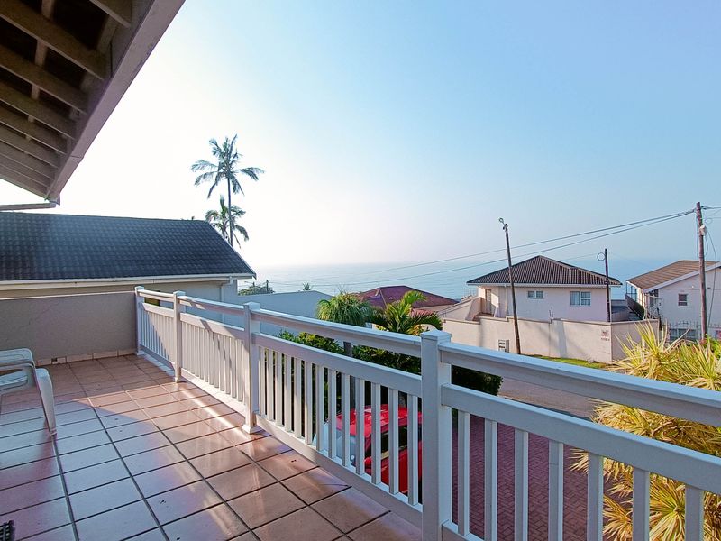 A spacious covered balcony featuring tiled flooring and a white balustrade railing, offering elevated views of the surrounding neighborhood and distant ocean.