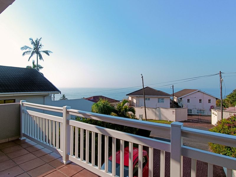 A covered balcony featuring tiled flooring and a white balustrade railing, offering an elevated view of the surrounding neighborhood and the ocean.