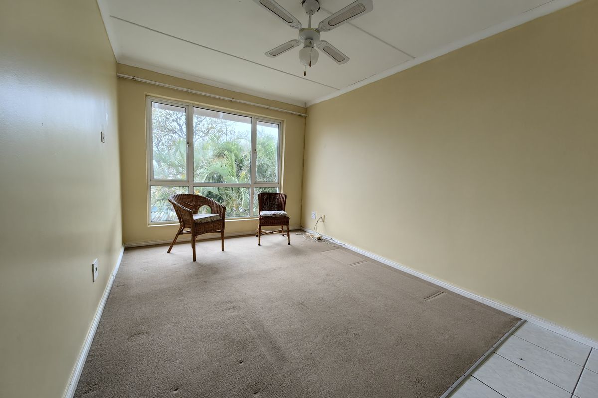 A spacious, empty living area featuring a ceiling fan, large window, and a combination of carpet and tiled flooring.