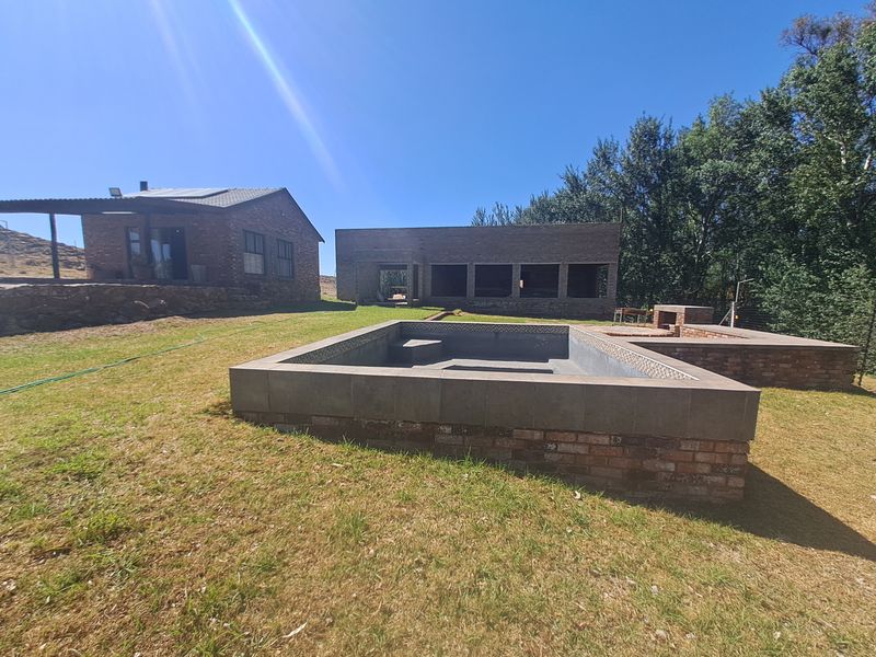 Outdoor area featuring a raised brick swimming pool and built-in braai, set against a backdrop of unfinished brick structures on a farm property.