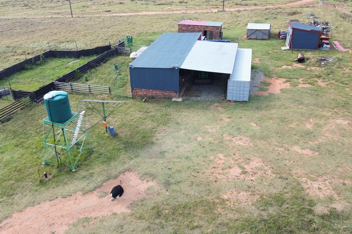 Aerial view of a farm property featuring multiple outbuildings, a water tank, a windmill, and fenced enclosures on open grazing land.