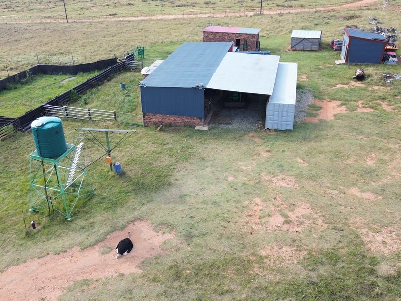 Aerial view of a farm property featuring multiple outbuildings, a water tank, a windmill, and fenced enclosures on open grazing land.