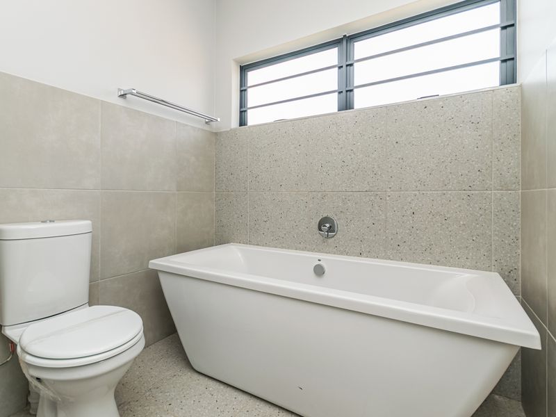 A modern, clean bathroom featuring a white freestanding bathtub, a toilet, and neutral-toned tiled walls and flooring with a horizontal window.