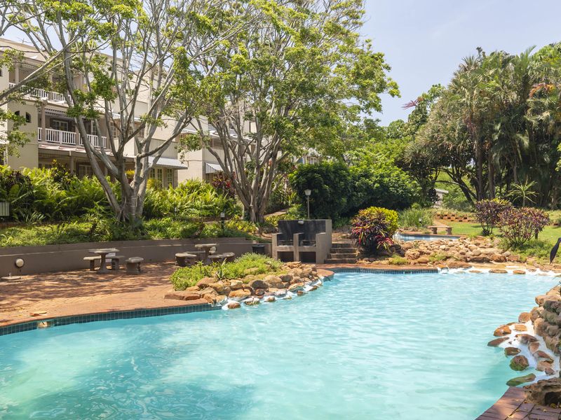 A well-maintained communal swimming pool area featuring a built-in braai, brick paving, and lush landscaped surroundings within the townhouse complex.