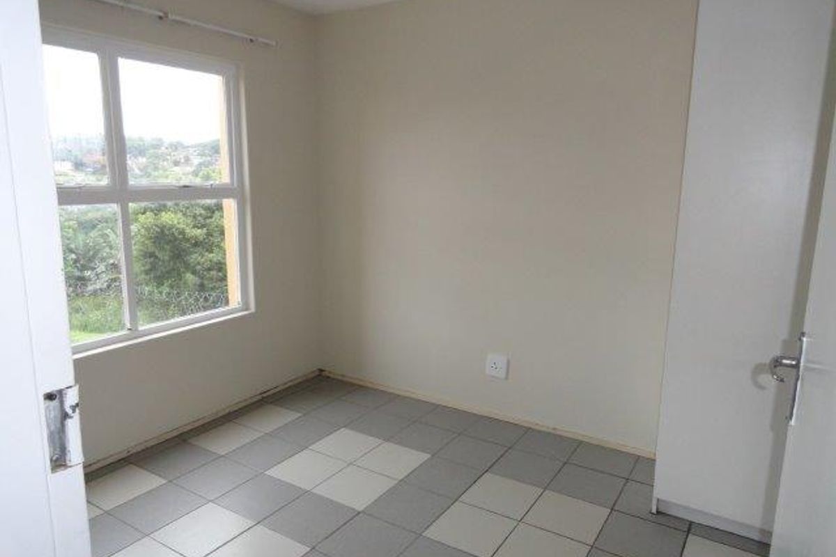 A clean, empty bedroom featuring tiled flooring, a built-in cupboard, and large windows providing natural light and a pleasant outlook.