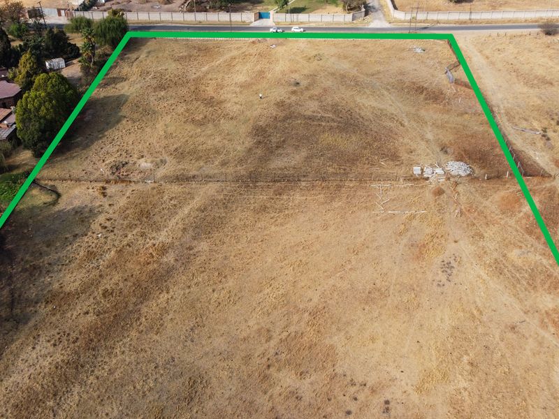 An aerial view of a vacant residential plot featuring dry grass, boundary fencing, and direct road access, suitable for future development.
