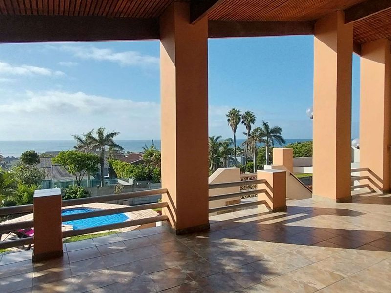 Spacious covered patio featuring tiled flooring, timber-slat ceiling, and structural pillars, offering elevated views over the garden, swimming pool, and distant ocean.