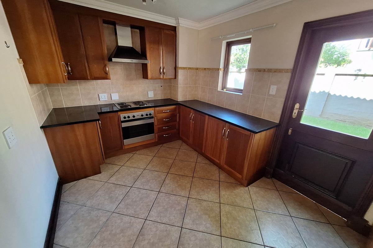A functional kitchen featuring warm-toned wooden cabinetry, black granite countertops, tiled flooring, and integrated appliances, with direct access to the exterior.