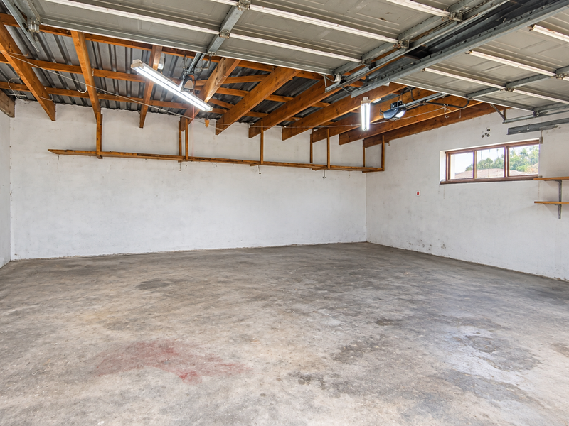 Spacious double garage featuring exposed timber roof trusses, concrete flooring, and wall-mounted shelving, providing ample storage and parking space.