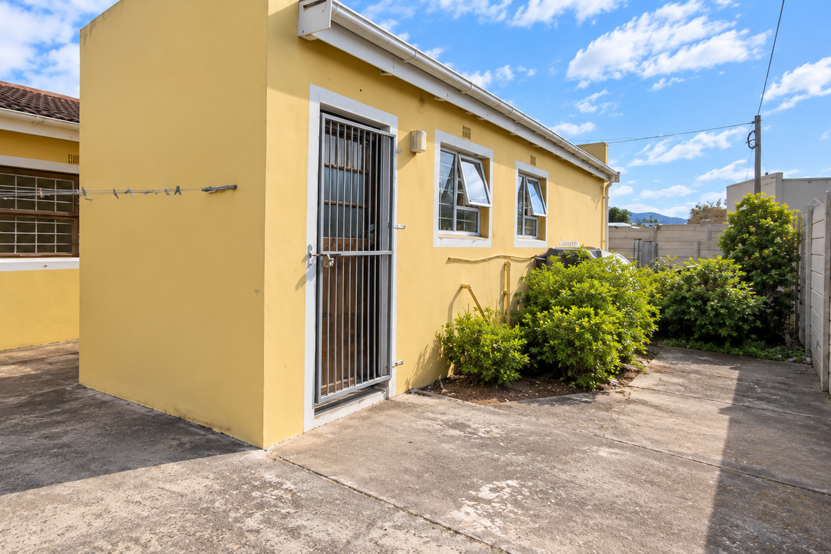 Rear exterior view of the property featuring yellow painted walls, security gate, casement windows, and a paved courtyard area with perimeter walling.