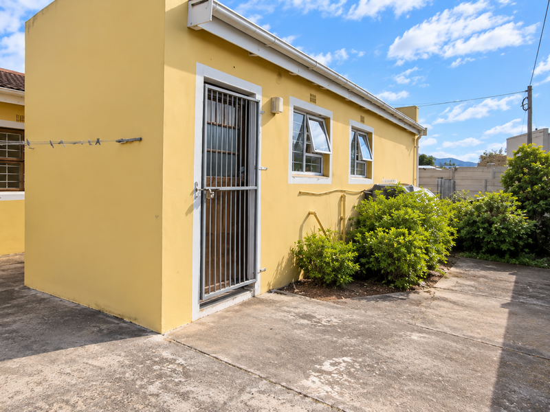 Rear exterior view of the property featuring yellow painted walls, security gate, casement windows, and a paved courtyard area with perimeter walling.
