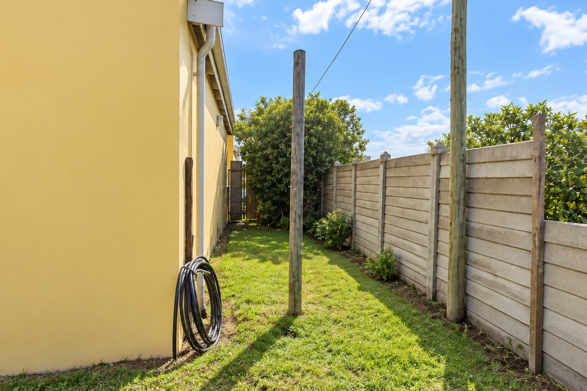 A side garden area featuring a yellow exterior wall, a precast concrete boundary fence, and established greenery.