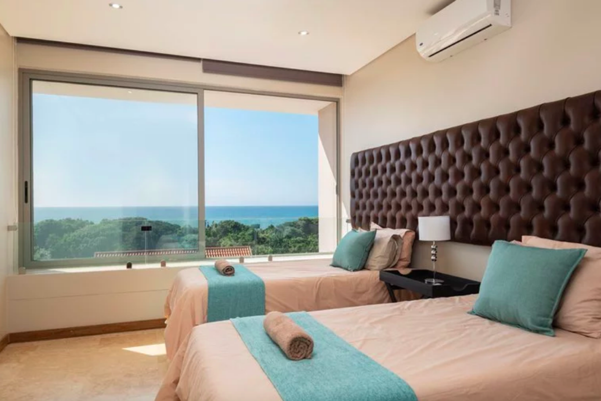 A modern bedroom featuring stone flooring, recessed lighting, a wall-mounted air conditioner, and a large sliding glass door opening to a scenic ocean view.