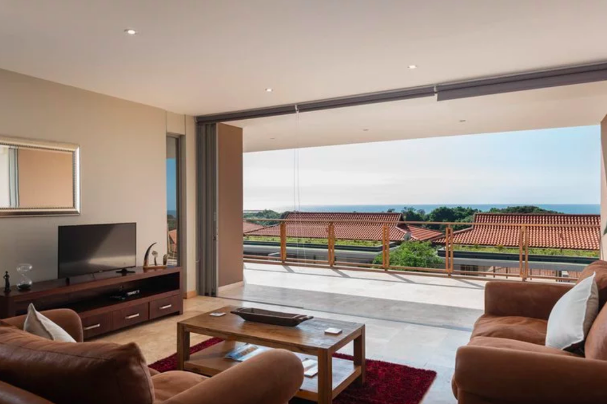 Spacious living room featuring large sliding glass doors that open onto a balcony with scenic coastal views and modern recessed lighting.