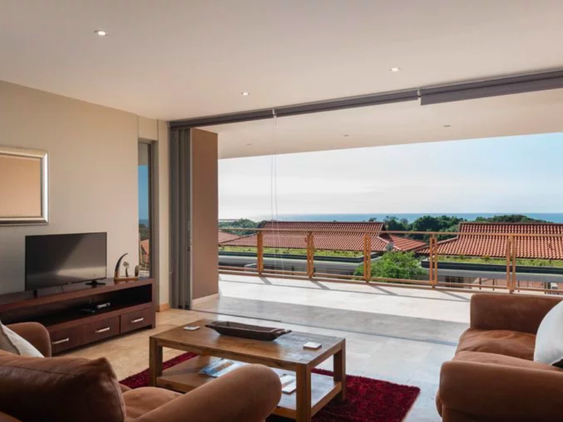 Spacious living room featuring large sliding glass doors that open onto a balcony with scenic coastal views and modern recessed lighting.