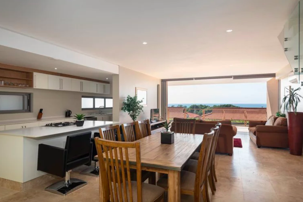Spacious open-plan living and dining area featuring a modern kitchen with stone countertops, gas cooktop, and seamless flow to a balcony with ocean views.