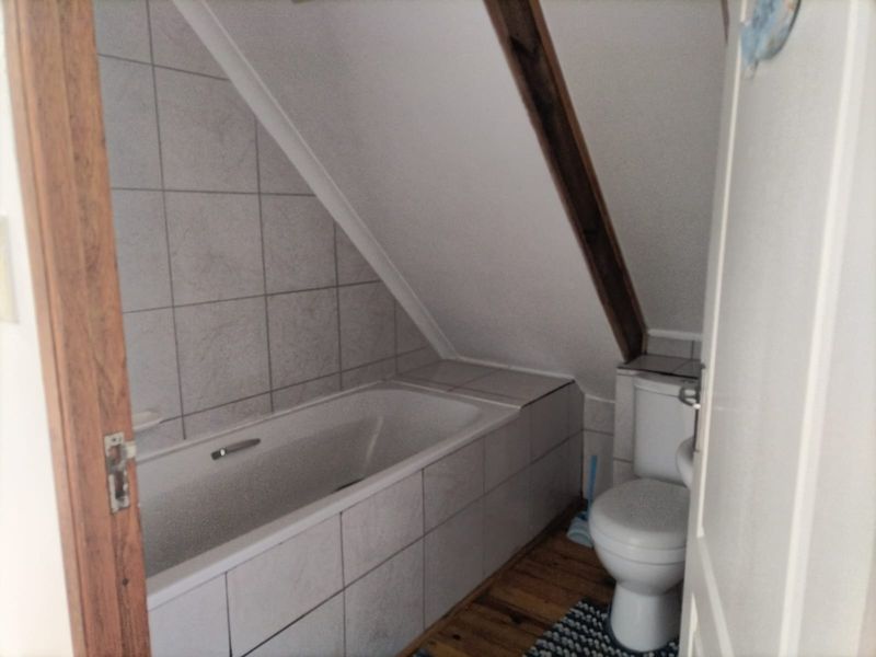 Compact attic-style bathroom featuring a built-in bathtub, toilet, tiled wall finishes, and timber flooring under a sloped ceiling.