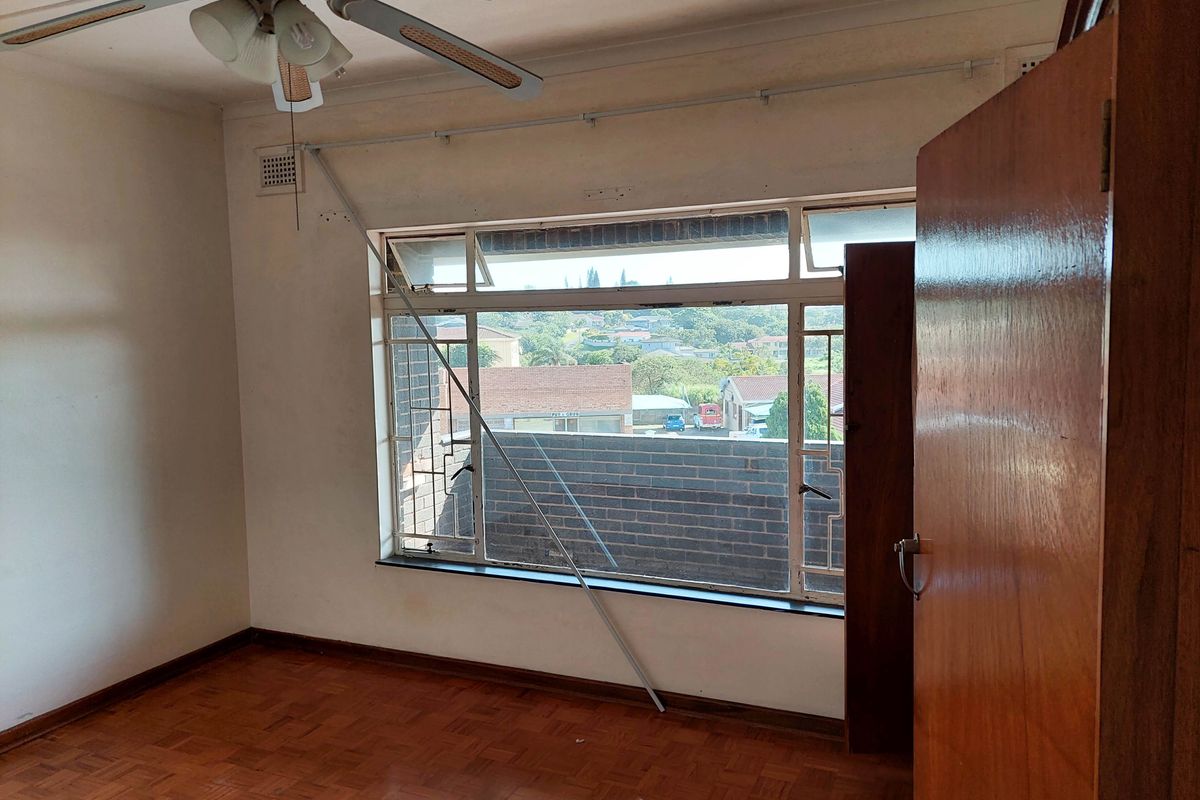 A bedroom featuring parquet flooring, a ceiling fan, and a large window with security bars, offering a view of the surrounding neighborhood.
