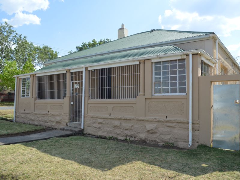 Front elevation of a single-story house featuring a corrugated metal roof, stone masonry base, and security bars on windows and entrance.