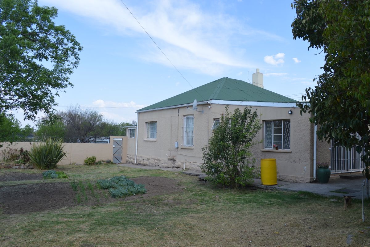 Rear view of a single-story house featuring a green metal roof, stucco walls, and security bars on the windows, set within a garden area.
