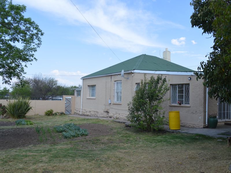 Rear view of a single-story house featuring a green metal roof, stucco walls, and security bars on the windows, set within a garden area.