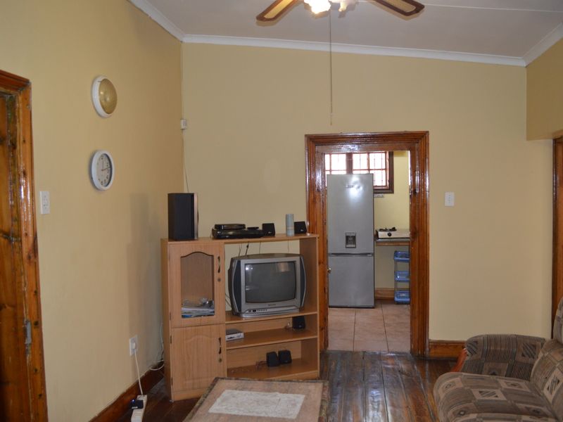 A living room featuring wooden flooring, traditional wooden door frames, and a ceiling fan, with a view into the adjacent kitchen area.