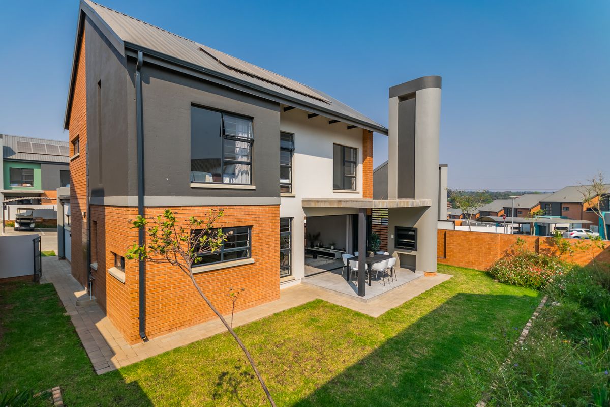 Rear view of a modern two-story house featuring a covered patio with a built-in braai, landscaped garden, and brick and rendered exterior finishes.
