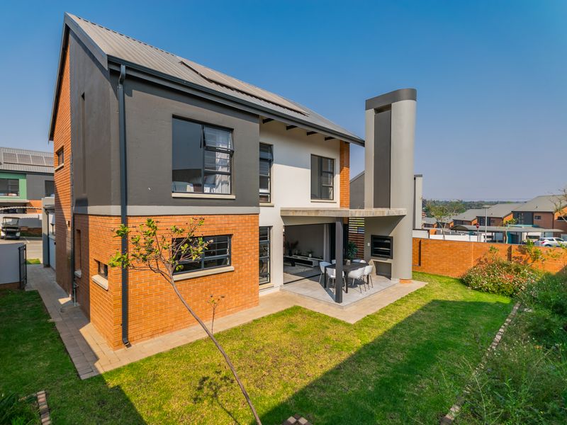 Rear view of a modern two-story house featuring a covered patio with a built-in braai, landscaped garden, and brick and rendered exterior finishes.