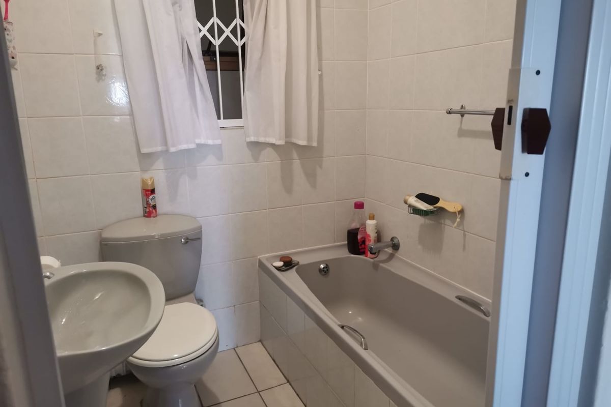 A functional bathroom featuring a bathtub, pedestal basin, and toilet, finished with neutral wall tiles and a security gate on the window.