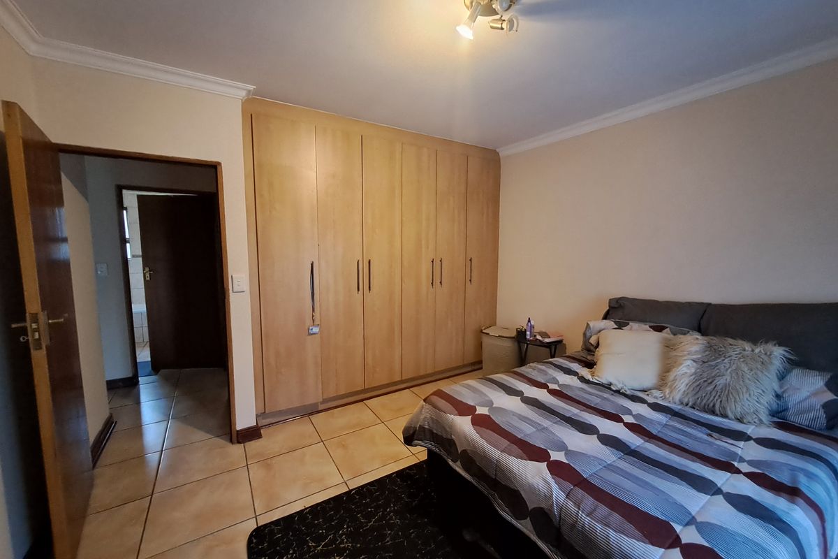 A bedroom featuring built-in wooden cupboards, tiled flooring, and a ceiling light fixture, with an adjacent hallway leading to another room.