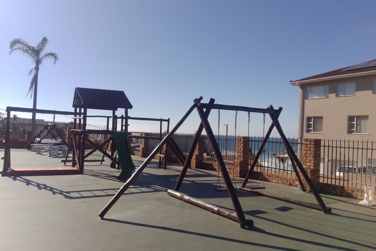 Outdoor communal play area featuring wooden swing sets and climbing structures, set on a paved surface with perimeter brick and metal fencing.