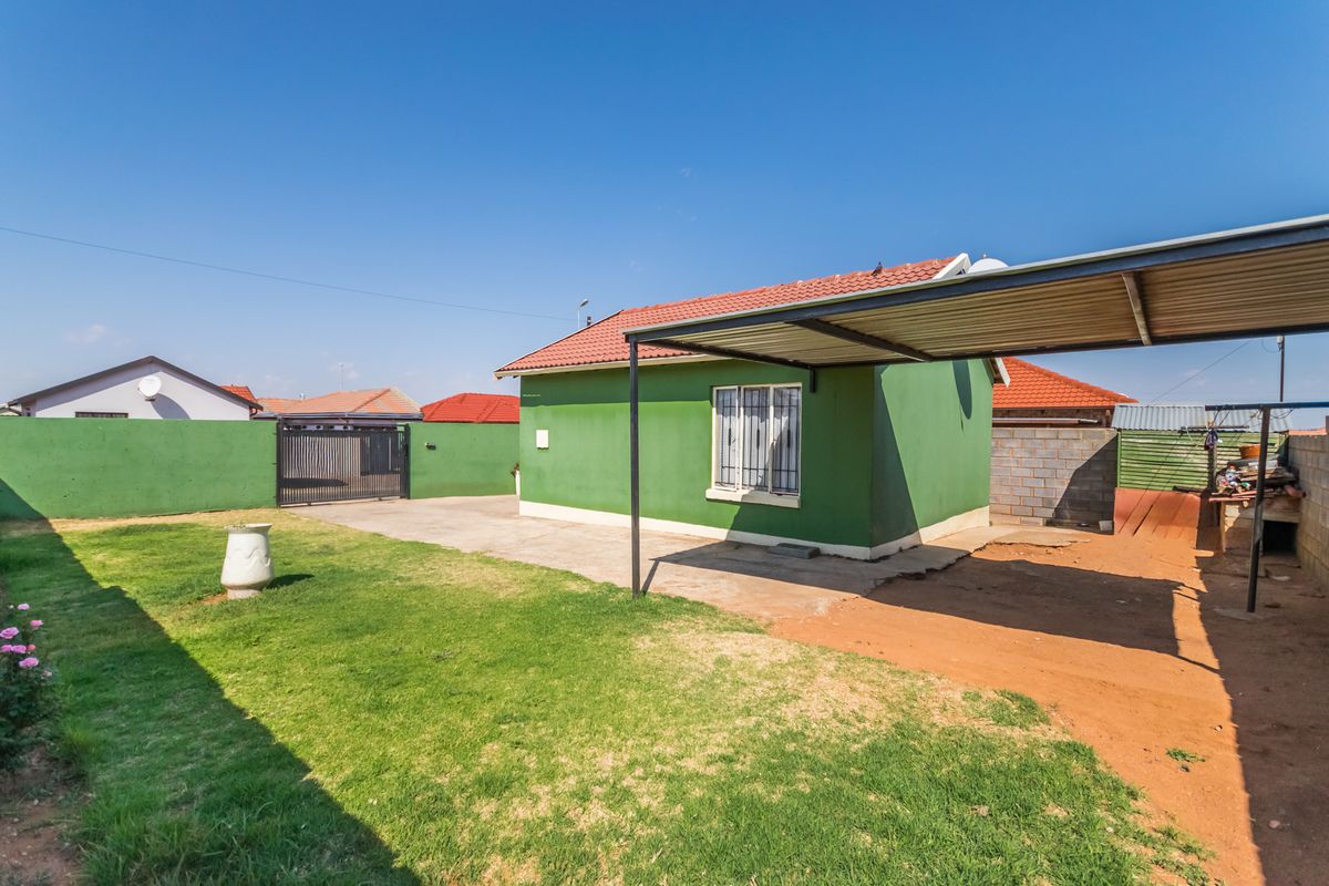 Rear view of the property featuring a green exterior facade, a metal carport, a lawn area, and a secure perimeter wall with a gate.