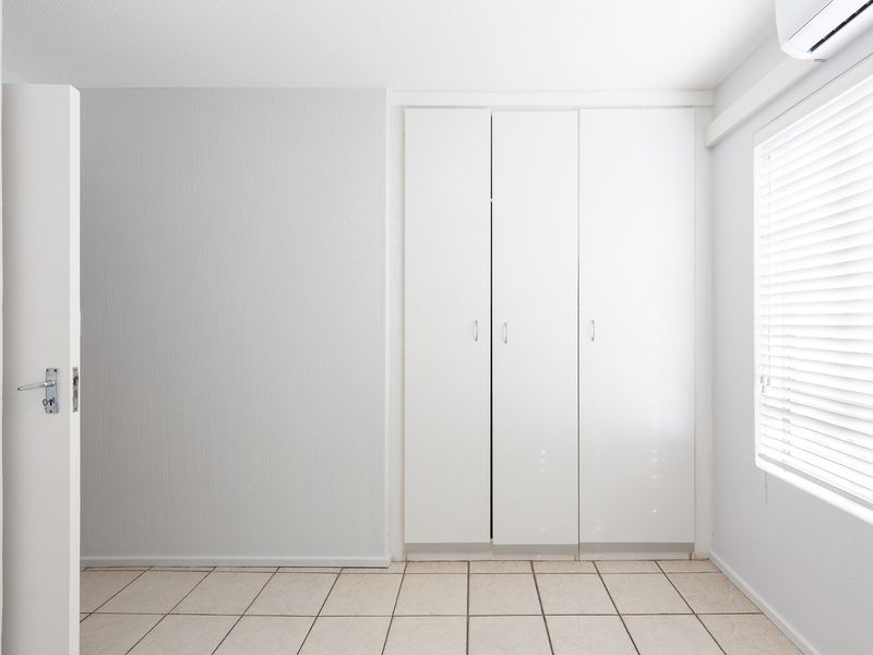 A bright, empty bedroom featuring neutral tiled flooring, built-in white cupboards, venetian blinds, and a wall-mounted air conditioning unit.