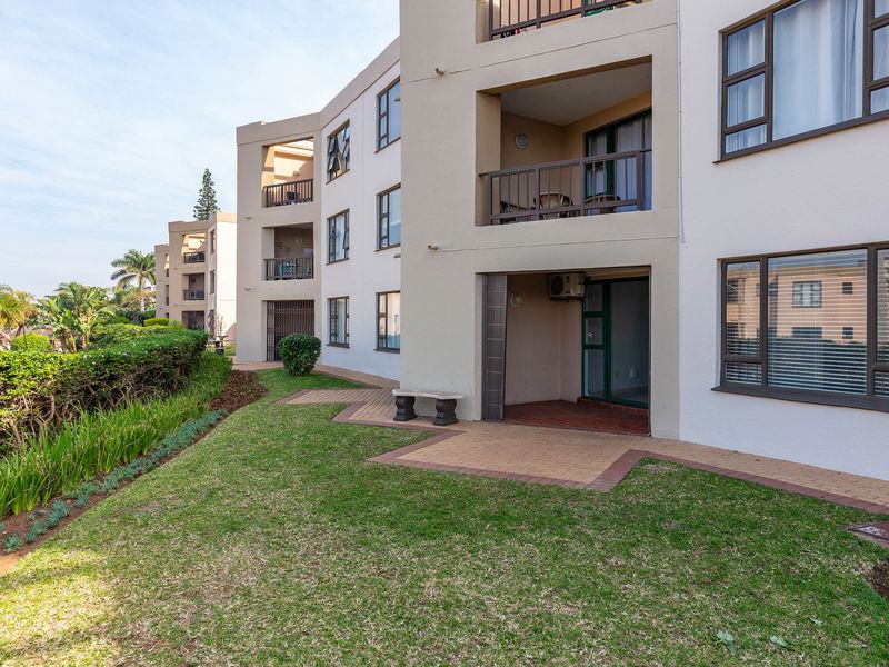 Rear view of the apartment complex featuring ground floor paved patios, landscaped garden areas, and upper-level balconies with aluminium window frames.