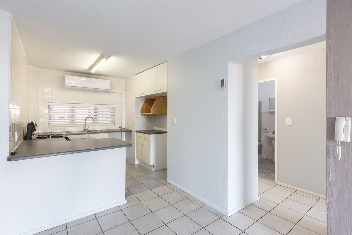 A functional kitchen featuring tiled flooring, ample cabinetry, laminate countertops, and a wall-mounted air conditioning unit, with an adjacent hallway leading to the bathroom.