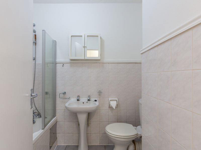 A functional bathroom featuring a pedestal sink, toilet, and a bathtub with a glass shower screen, finished with neutral wall and floor tiles.