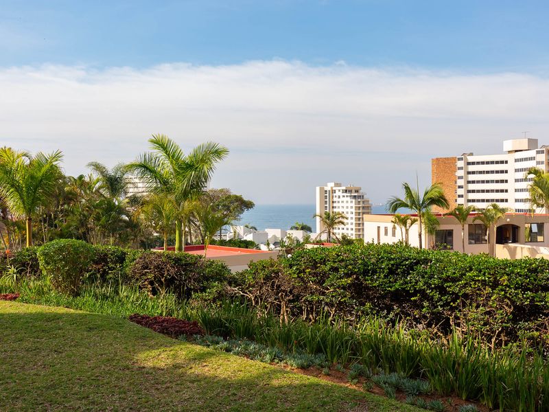 A scenic elevated view overlooking a landscaped garden with lush greenery, palm trees, and distant coastal buildings under a clear blue sky.