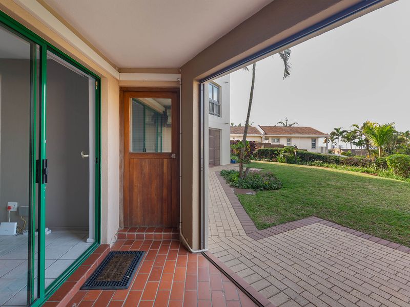A covered patio area with terracotta tiling, featuring a sliding glass door and direct access to a paved walkway and communal garden.